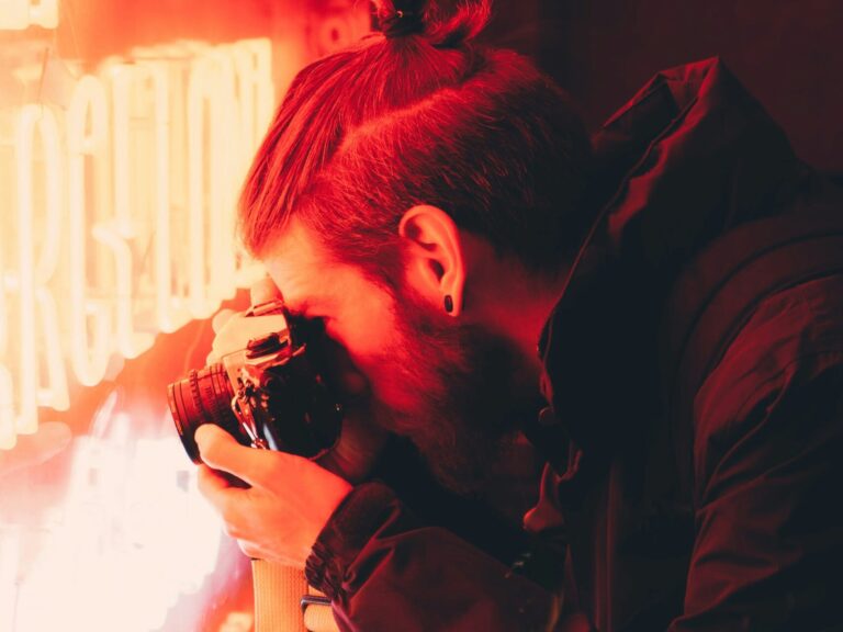 Ein Mann Fotografiert ein rotes Leuchtschild, das Bild ist dunkel und der Mann trägt eine schwarze Jacke